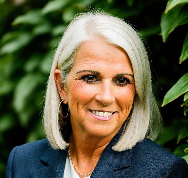A photo of LaDonna Williams, a white woman with blonde shoulder length hair. She is posing for a photo outside in front of a tree while looking past the camera as if in thought.