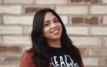 Photo of Genesis. A young woman with brown skin and long dark hair. She is smiling and posing for a photo in front of a brick wall that is slightly out of focus.