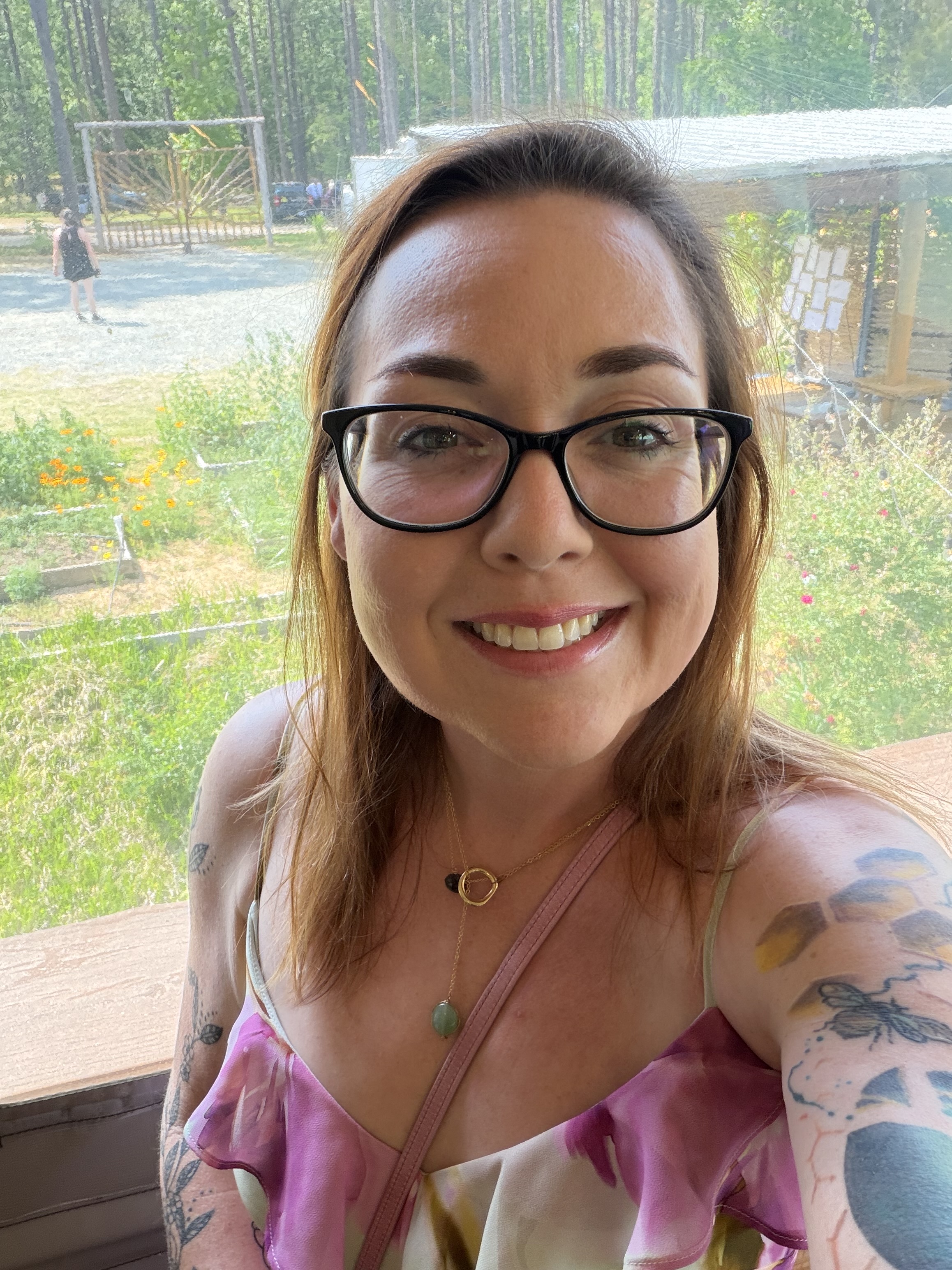 Photo of Rachel Kaplan. A white woman in her late thirties with dark rimmed glasses and long reddish hair. She is taking a selfie with a floral dress on with an urban garden in the background.