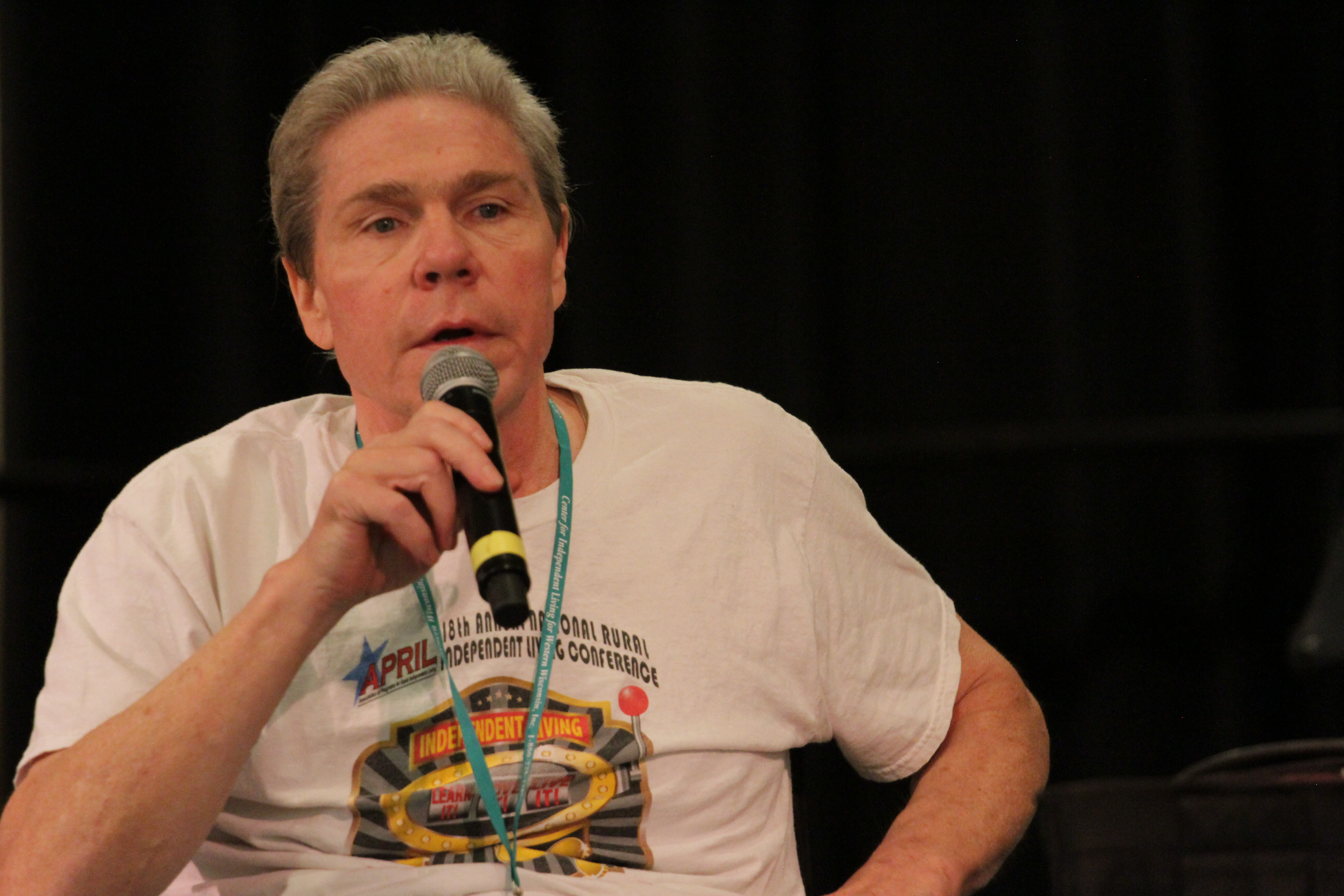 Photo of Tim Sheehan, an older white man with close cut salt and pepper hair wearing a white APRIL conference t-shirt and speaking into a microphone.